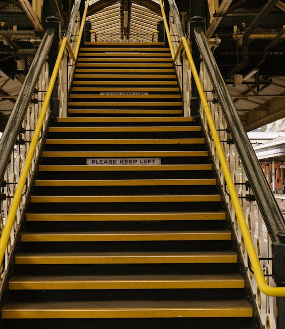 A straight staircase inside an industrial or warehouse setting, with metal railings painted yellow on both sides and steps covered with a dark anti-slip surface. The staircase has safety signs on the